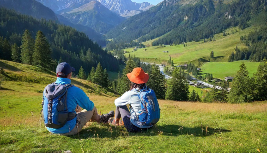 A couple sitting at the summit of a mountain trail with backpacks, gazing over a vast valley surrounded by mountains, with dandelion seeds floating in the breeze, symbolizing resilience, accomplishment, and breathing easy in the fight against lung cancer.